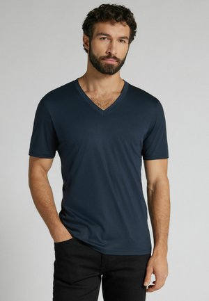 Man met baard en krullend haar, gekleed in een donkerblauw V-hals T-shirt met korte mouwen en een zwarte broek, staand tegen een effen achtergrond.