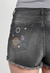Pantalones cortos de mezclilla gris con dobladillo deshilachado, que presentan bordados con temática espacial en el bolsillo trasero, incluyendo un planeta, estrellas y un cohete.