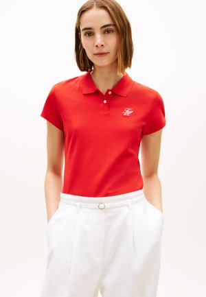 Tânără cu păr scurt și castaniu, purtând un tricou polo roșu cu logo alb și pantaloni albi cu talie înaltă și curea subțire, mâinile în buzunare.