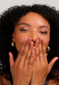Kvinna med lockigt hår och guldfärgade örhängen täcker munnen med händerna, visar ringar och välpolerade naglar i naturfärg mot en enfärgad bakgrund.