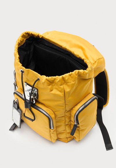 Sac à dos en tissu jaune avec deux poches avant zippées, intérieur noir, fermeture par cordon et sangle d'épaule noire réglable sur fond clair.