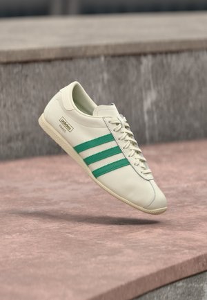 Baskets blanches Adidas avec des rayures vertes et le texte « PARIS », flottant au-dessus d'une surface en pierre avec un mur gris en arrière-plan.