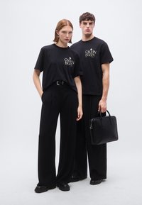 Μαύρα βαμβακερά μπλουζάκια με λογότυπο, σε συνδυασμό με μαύρο παντελόνι φαρδιάς γραμμής. Κάποιος κρατά μια μαύρη δερμάτινη τσάντα, με μινιμαλιστικό σχεδιασμό και μεταλλικά στοιχεία.