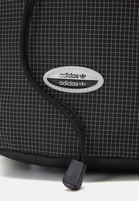 Black backpack with a grid pattern, featuring a grey oval logo with "adidas" in black. Includes a black zipper pull and textured fabric.