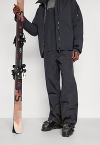 Άτομο που φοράει μαύρη στολή σκι και μπότες, κρατώντας μια όρθια ζεύγαρι καφέ και μαύρες σανίδες σκι με δέστρες σε ουδέτερο φόντο.