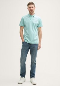 Hellgrünes Polohemd mit klassischem Kragen und einer Knopfleiste mit drei Knöpfen, kombiniert mit blauen Jeans und weißen Turnschuhen.
