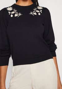 Pull noir en tricot avec manches bouffantes et broderie florale sur les épaules. Présente des poignets et un ourlet côtelés, texture du tissu visible.