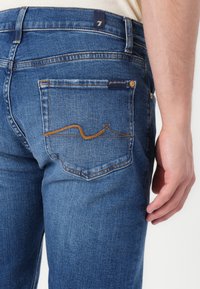 Sinised denim püksid, mis on sirge lõikega, pruuni tikandiga disain tagakotil ja väike silt vöökohtades.