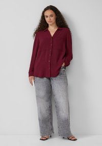 Bordeaux knoop-overhemd met twee borstzakken, gecombineerd met losse grijze jeans. Het overhemd heeft een lichte textuur en een kraagontwerp. Zwarte hoge hak sandalen.