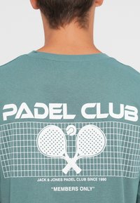 Turquoise T-shirt featuring "PADEL CLUB" in white, with two crossed padel rackets against a grid pattern. Text details include "MEMBERS ONLY."