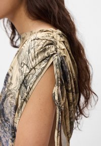 Beige top met abstract zwart en grijs patroon, textuurstof, gedrapeerd ontwerp en strikken op de schouders; open arm details zichtbaar.