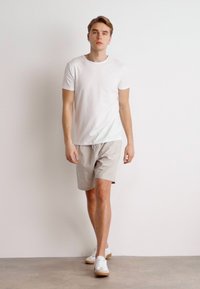 Hvid kortærmet t-shirt parret med lysebeige shorts. Model iført hvide sneakers, står foran en neutral baggrund.