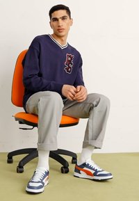 Jūras spēku džemperis ar baltu apmalīti un sarkaniem uzrakstiem, gaiši pelēkas bikses un zilas sporta apavus ar oranžiem akcentiem. Modelis sēž uz oranžas krēsla.