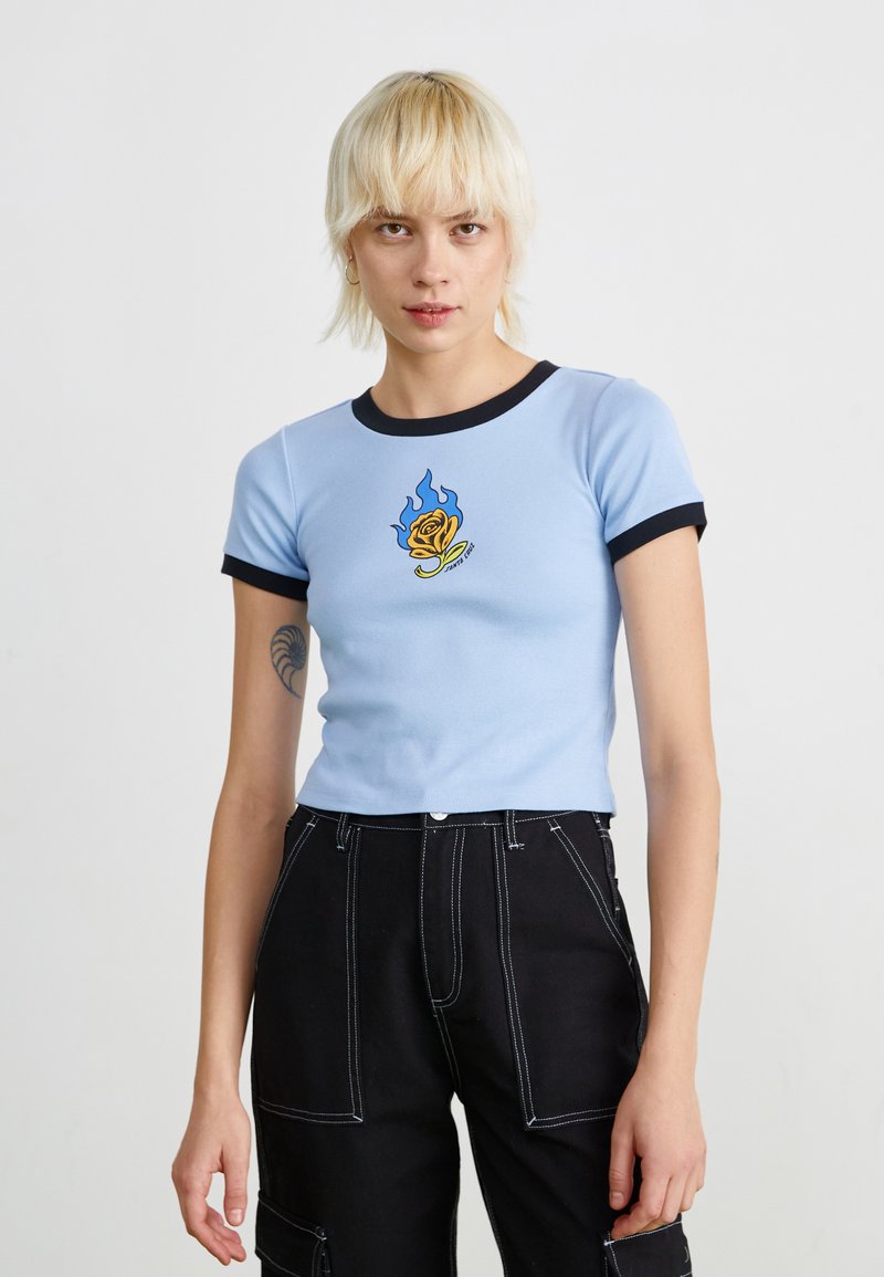 Top azzurro chiaro a maniche corte con bordi neri, presenta una grafica di una rosa e fiamme al centro. Indossato con pantaloni cargo neri.