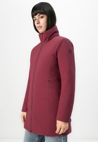 Vrouw in een wijnrode winterjas met ritssluiting en hoge kraag, staand tegen een effen witte achtergrond.