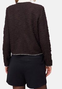 Pull court texturé noir avec manches longues et un accent argenté à l'ourlet. Porté sur un short foncé, mettant en valeur un design superposé.