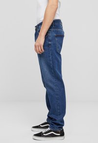 Jeans de mezclilla azul claro con un corte relajado, cinco bolsillos y costuras visibles. Combinados con zapatillas bajas negras y blancas.