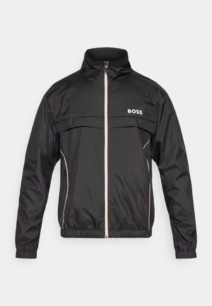 Chaqueta cortavientos negra con cuello alto, cremallera frontal, logo BOSS en el pecho y detalles en beige contrastante a lo largo de la cremallera y los bolsillos.