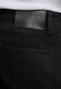 Detailný záber na zadnú kapsu čiernych džínsov, ktorá zobrazuje prešitú koženú nálepku so slovami "AY' STUDIOS" vyrazenými nad kapsou.