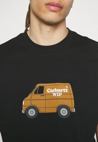 Hombre con una camiseta negra con un gráfico de una furgoneta marrón y el logo blanco de "Carhartt WIP" en el pecho, frente a un fondo liso.