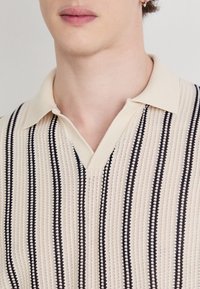 Beżowa, dziana koszulka polo z dekoltem w serek, pionowymi czarnymi paskami, teksturowanym materiałem oraz klasycznym kołnierzykiem.