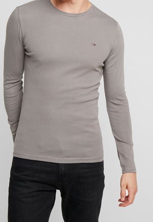 Man wearing a fitted gray long-sleeve shirt and black jeans, standing against a plain white background.