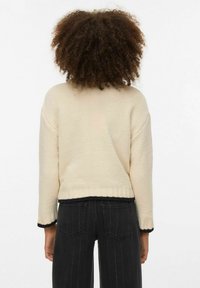 Suéter de punto color crema con acabado texturizado, diseño crop y ribete negro a lo largo del dobladillo y las mangas. Se lleva sobre pantalones oscuros con rayas.