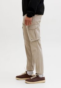 Person trägt beige Cargo-Hosen mit großen Seitentaschen, weiße Socken, braune Sneaker mit niedrigem Schaft und eine schwarze Jacke, gezeigt von der Taille abwärts.