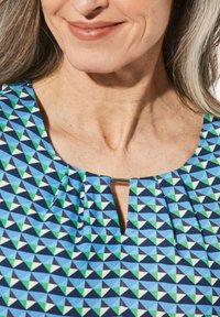 Top blu con un motivo geometrico in verde, bianco e blu navy. Presenta un piccolo foro a chiave sul collo e delle pieghe.