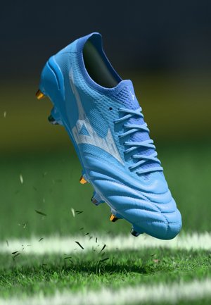 Cleat de fotbal albastru deschis cu logo alb și șireturi, suspendat în aer deasupra unui teren de iarbă cu o linie albă.