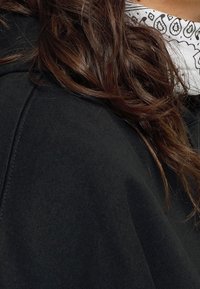 Sudadera de tela negra con una textura suave, que muestra una parte de largo cabello castaño ondulado y una bandana blanca con patrones negros debajo.