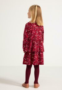 Czerwona sukienka w kwiatowy wzór z warstwowym krojem, długimi rękawami, zestawiona z bordowymi rajstopami i brązowymi butami. Dziecko widziane z tyłu.