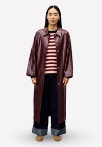 Frau, die einen langen glänzenden burgunderfarbenen Mantel über einem rosa-rot gestreiften Pullover, weiten, gekrempelten Jeans und beigen Schuhen trägt, aufrecht steht.