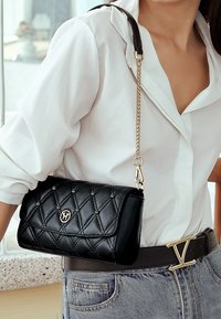 Gequilted zwarte leren handtas met gouden accenten, kettingriem en logo. De tas wordt crossbody gedragen, gecombineerd met een witte shirt en denim.