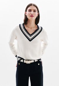 Maglione crema a collo a V con accentuazioni a righe blu navy, texture lavorata a maglia, abbinato a pantaloni scuri e una cintura bianca con una fibbia dorata.