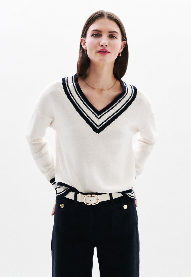 Maglione crema a collo a V con accentuazioni a righe blu navy, texture lavorata a maglia, abbinato a pantaloni scuri e una cintura bianca con una fibbia dorata.