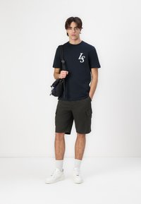 T-shirt bleu marine à col ras du cou avec logo "LS" blanc, shorts cargo olive, baskets blanches et une sacoche noire. Fond uni.