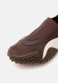 Gros plan d'une chaussure marron sans lacets avec une semelle en caoutchouc texturée et des détails blancs sur les côtés, fabriquée en tissu et en cuir.