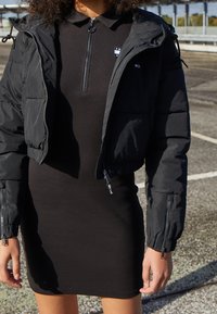 Czarna kurtka puchowa z zamkiem z przodu i elastycznymi mankietami na dopasowanej czarnej sukience. Sukienka ma kołnierzyk i zamykany na zamek przód.