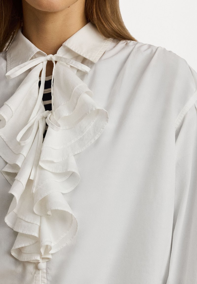 Biała bluzka z falbaniastym przodem o warstwowej, miękkiej teksturze, z wiązaną kokardą przy dekolcie i klasycznym wzorem kołnierzyka.