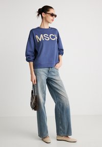 Μπλε φούτερ με χρυσά γράμματα "MSCH", συνδυασμένο με ανοιχτό μπλε παντελόνι φαρδύ και ανοιχτά παπούτσια. Το μοντέλο κρατά μια σκούρα τσάντα.