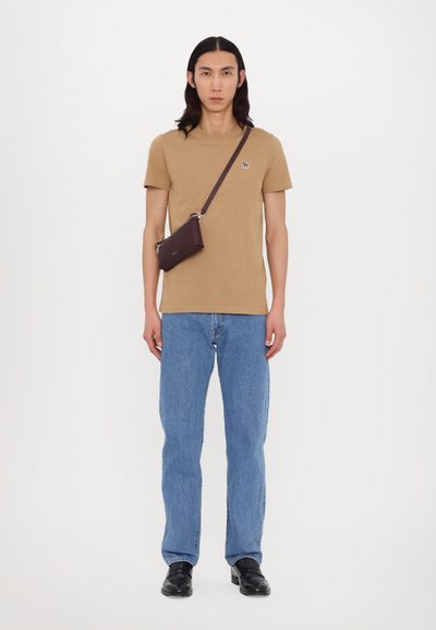Tan t-shirt with a small logo, blue denim jeans, and black shoes. A small dark brown crossbody bag is worn across the torso.