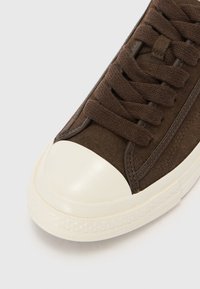 Hnědé semišové tenisky s krémovou gumovou špičkou, texturovanými tkaničkami a kontrastním tmavším prošíváním po stranách.