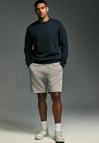 Mörk marinblå Crewneck tröja, ljus beige shorts, vita strumpor och grå sneakers med en texturerad sula, stående mot en neutral bakgrund.