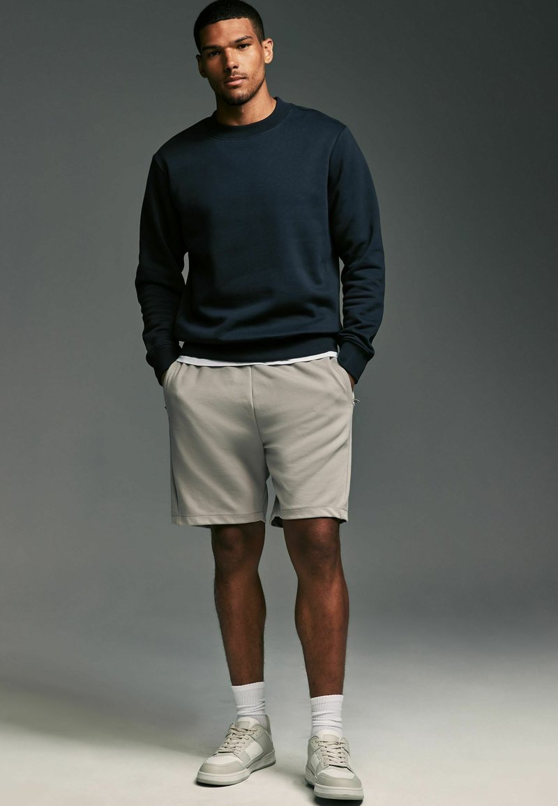 Mörk marinblå Crewneck tröja, ljus beige shorts, vita strumpor och grå sneakers med en texturerad sula, stående mot en neutral bakgrund.