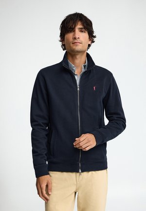 Hombre vestido con chaqueta azul marino con cremallera sobre una camisa azul claro y pantalones beige, de pie sobre un fondo blanco liso.
