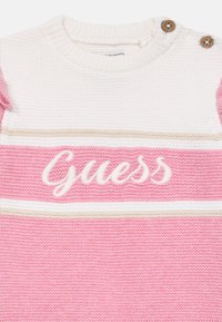 Rózsaszín kötött pulóver fehér és bézs csíkokkal, melyen fehér hímzéssel szerepel a "Guess" felirat. Két fa gomb található a vállán.