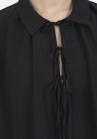 Zwarte blouse met een kraag, voorzien van een keyhole-halslijn en een strikdetail. Zachte textuur, losse pasvorm en vloeiende stof zonder extra patronen.
