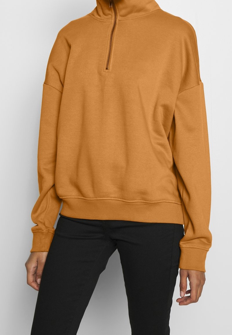 Femme portant un pull léger marron clair avec une courte fermeture éclair au niveau du cou, associée à un pantalon noir, sur un fond uni.