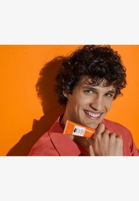 En oransje hudpleietube holdes av en person, med en hvit etikett med svart tekst. Bakgrunnen er en lys oransje vegg. Personen har på seg en korallfarget skjorte.